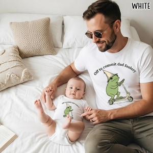 May include: White t-shirts and onesie with a green frog graphic riding a scooter and the text "Off to commit dad foolery". The adult is wearing sunglasses and the baby is lying on a white bed.
