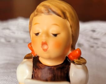May include: A ceramic figurine of a seated child singing from a sheet of music. The figurine has blonde hair with orange bows, a brown dress, and white sleeves. The child's cheeks are rosy, and the figurine is on a white surface.