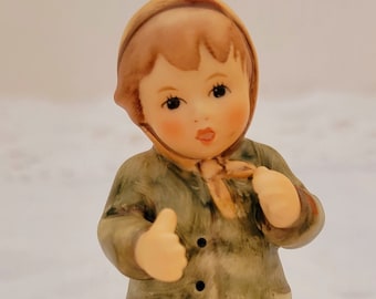 May include: A porcelain figurine of a young girl wearing a green coat, brown boots, and a brown hat with a red trim. She is holding her hands up in a gesture of greeting.