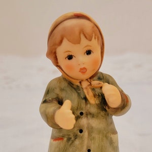 May include: A porcelain figurine of a young girl wearing a green coat, brown boots, and a brown hat with a red trim. She is holding her hands up in a gesture of greeting.