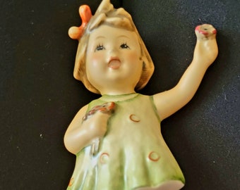 May include: A porcelain figurine of a girl wearing a green dress with white polka dots and brown shoes. She is holding a bouquet of flowers and has a red bow in her hair.