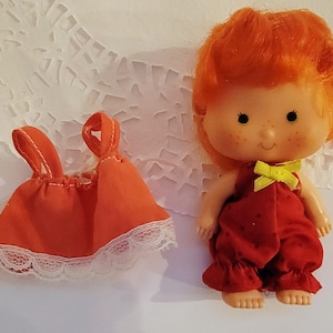 May include: A small doll with bright orange hair, freckles, and a red romper featuring a yellow bow at the neckline. Beside the doll is an orange sleeveless top with white lace trim along the bottom hem, both made of fabric materials.
