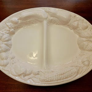 May include: An oval-shaped, cream-colored ceramic serving platter divided into two sections. The rim is decorated with a raised, embossed relief pattern featuring various harvest vegetables, including pumpkins, squash, corn, and acorns.