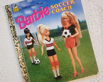May include: A Little Golden Book titled Barbie Soccer Coach featuring an illustration of three dolls on a soccer field. The cover shows dolls in white and black athletic uniforms with soccer balls, set against a green grass background.