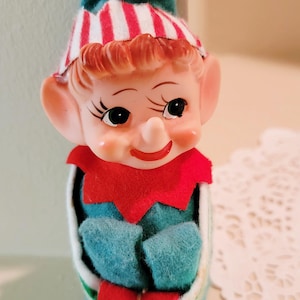 May include: A vintage Christmas elf ornament. The elf has a painted face with rosy cheeks, a red collar, and a teal outfit. The elf's hat is teal with red and white stripes. A small gold bell hangs from the hat.