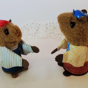 May include: Two flocked squirrel figurines facing each other. The left squirrel wears a red cap and a blue and white striped shirt. The right squirrel wears a blue cap, a yellow patterned shirt with a blue tie, and red striped trousers.