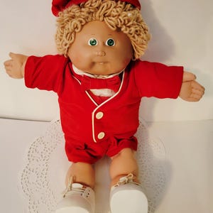 May include: A vintage Cabbage Patch Kid doll with blonde yarn hair, green eyes, and a red outfit. The doll is wearing a red hat, jacket, shorts, and bow tie. White shoes complete the ensemble. The doll is posed with arms outstretched.
