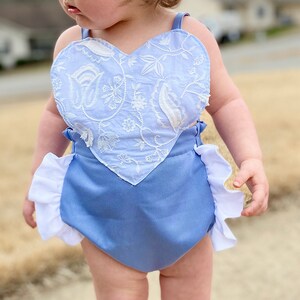 Puede incluir: Una niña pequeña con un mameluco azul claro con un diseño bordado en forma de corazón blanco. El mameluco tiene volantes blancos alrededor de las piernas.