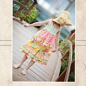 May include: A young girl wearing a colorful floral dress with ruffles and a straw hat with a flower accent. The dress has a pink, yellow, and green floral pattern. The girl is standing on a wooden bridge. The image is from CKC Patterns, Couture Made Simple, CKC Kids.