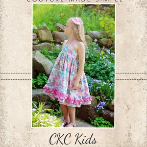 May include: A young girl wearing a pink and blue floral dress with a pink ruffle trim stands on a stone path in a garden. The dress has a halter neckline and is sleeveless. The girl is looking to the right of the image. The image is from CKC Patterns, Couture Made Simple, CKC Kids.