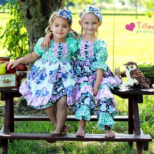 May include: Two young girls wearing floral print dresses with ruffles sit on a wooden picnic table. The girls are smiling and have their arms around each other. The table is decorated with apples and a wooden crate that says "Harvest Apples".