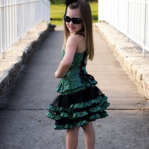 May include: A young girl wearing a green and black leopard print dress with black ruffles and a black bow. She is wearing black sunglasses and a black feather headband. She is standing on a paved path with a white picket fence in the background.