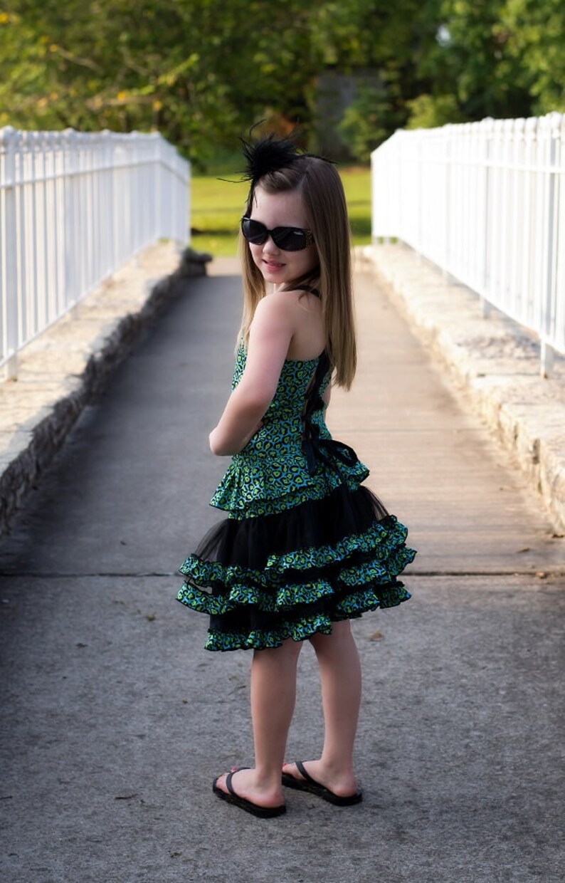 May include: A young girl wearing a green and black leopard print dress with black ruffles and a black bow. She is wearing black sunglasses and a black feather headband. She is standing on a paved path with a white picket fence in the background.