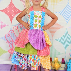 May include: A young girl wearing a colorful layered dress with a ruffled bodice and a white background with birds and turtles. The dress has a variety of patterns including polka dots, flowers, and geometric shapes. The girl is standing in front of a colorful patchwork background.
