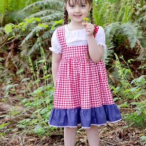 May include: A young girl wearing a red and white gingham dress with a blue denim ruffle trim. She is holding a yellow flower and has her hair in braids.