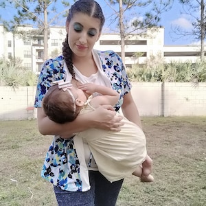 May include: A woman in a blue and white floral print shirt is holding a baby in a cream-colored onesie. The woman is wearing blue jeans and the baby is wearing a white headband with a blue bow.