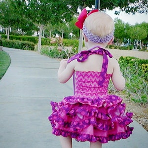 May include: A young girl wearing a pink and purple ruffled dress with a purple headband and a red bow. She is walking on a sidewalk with a green grassy area in the background.
