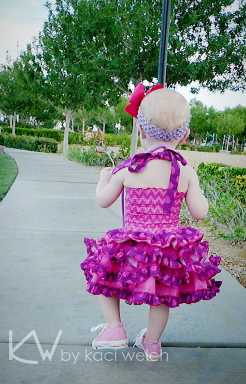 May include: A young girl wearing a pink and purple ruffled dress with a purple headband and a red bow. She is walking on a sidewalk with a green grassy area in the background.
