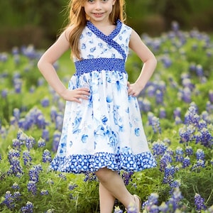 May include: A young girl wearing a white and blue floral dress with a blue and white patterned waistband and a blue and white ruffled hem. She is standing in a field of bluebonnet flowers.