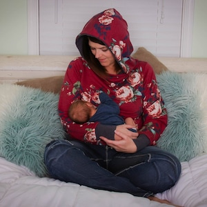 Könnte beinhalten: Eine Frau in einem roten Kapuzenpullover mit Blumenmuster sitzt auf einem Bett und hält ein Baby in ihren Armen. Das Baby trägt ein blaues Hemd und schläft.