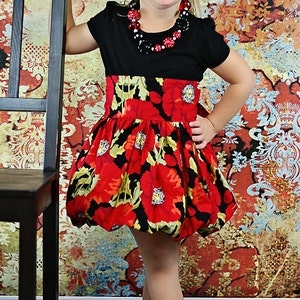 May include: A young girl wearing a black and red dress with a floral print. She is standing next to a black chair and smiling. She is wearing a black necklace with red and green beads.