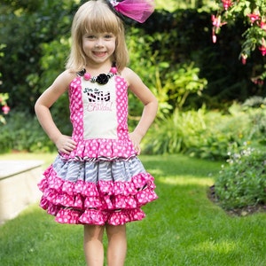 May include: A young girl wearing a pink and white polka dot dress with a zebra print ruffle and a black top hat with a pink tulle bow. The dress has the text "I'm a Wild Child!" embroidered on the front. She is standing in a grassy area with green foliage in the background.