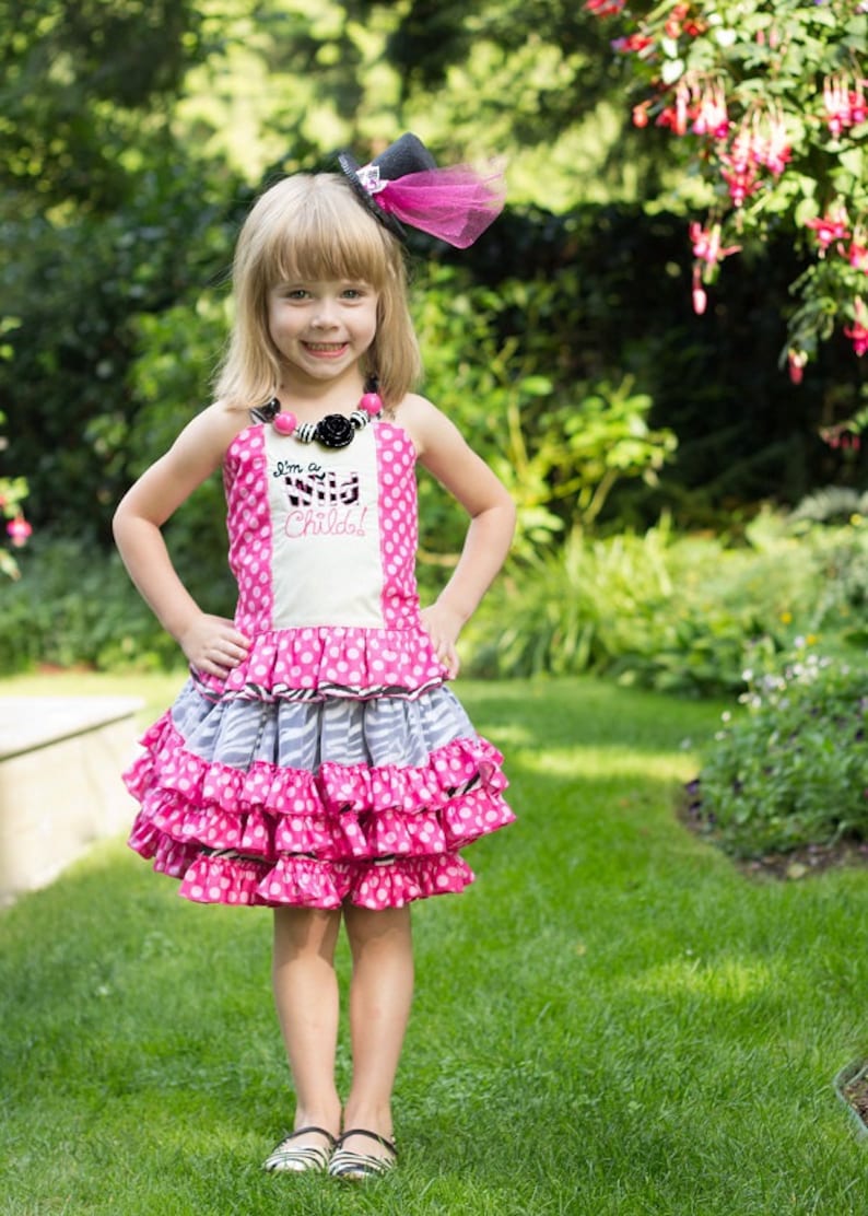 May include: A young girl wearing a pink and white polka dot dress with a zebra print ruffle and a black top hat with a pink tulle bow. The dress has the text "I'm a Wild Child!" embroidered on the front. She is standing in a grassy area with green foliage in the background.