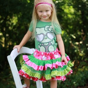 May include: A young girl wearing a green t-shirt with a Hello Kitty graphic and a pink headband. She is wearing a multi-layered skirt with pink, green, and white ruffles. She is standing on a white wooden chair with her feet on a wooden surface.