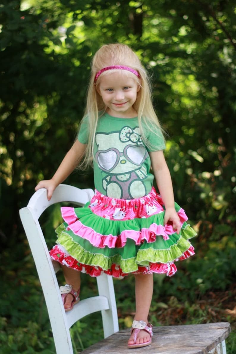May include: A young girl wearing a green t-shirt with a Hello Kitty graphic and a pink headband. She is wearing a multi-layered skirt with pink, green, and white ruffles. She is standing on a white wooden chair with her feet on a wooden surface.