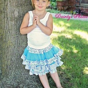 May include: A young girl wearing a white tank top and a blue tulle skirt with white floral accents. She is wearing a pink sequined headband and has her hands clasped in front of her. She is standing next to a tree.