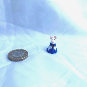 Könnte beinhalten: Eine Miniaturfigur einer Maus in einem blau-weißen Outfit mit rosa Ohren und einem weißen Gesicht. Neben der Figur liegt eine Münze. Der Hintergrund ist ein hellblauer Stoff.