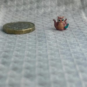 May include: A miniature brown owl-shaped teapot figurine with a small teal handle, positioned next to a gold-colored coin for scale. The owl has large dark eyes and a rounded body, showcasing intricate details in a compact size.