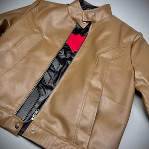 May include: A tan leather jacket with a black and red design on the chest. The jacket has a zipper closure and snap buttons. The collar is a stand-up style. The jacket is a stylish outerwear piece.