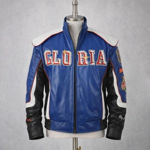 Op de afbeelding: Een blauwe leren jas met witte en zwarte accenten. De jas heeft het woord "GLORIA" in rode en witte letters over de borst. Het heeft een opstaande kraag en een ritssluiting over de volledige lengte. De mouwen hebben patches en tekst.