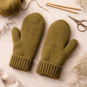 May include: A pair of olive green knitted mittens with ribbed cuffs. The mittens are made of yarn and are displayed with knitting needles, scissors, and a ball of yarn, suggesting a handmade craft project.