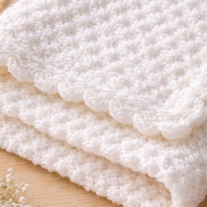May include: A folded, white, crocheted blanket with a scalloped edge. The blanket has a textured, raised pattern and is made of soft yarn. The blanket is folded in thirds, showing the texture and detail of the crochet work.