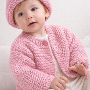 May include: A baby in a knitted pink hat and cardigan set. The cardigan has two large pink buttons. The baby is also wearing white trousers. The knitted set is made of a soft, textured yarn.