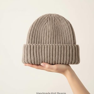 May include: A taupe-colored, ribbed knit beanie hat. The hat is held in a hand and has a folded brim. The text "Handmade Knit Beanie" is at the bottom of the image. This warm hat is perfect for cold weather.