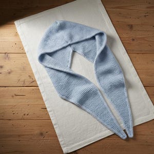 Pode incluir: Um conjunto de capuz e cachecol de malha azul claro. O capuz está conectado às pontas longas do cachecol, tudo sobre um pano de linho branco. O fundo é uma superfície de madeira.