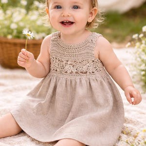 Puede incluir: Una niña sonriente con un vestido de top de ganchillo beige con una diadema a juego, sosteniendo una margarita. La niña está sentada en una manta de color crema al aire libre, rodeada de flores y vegetación.