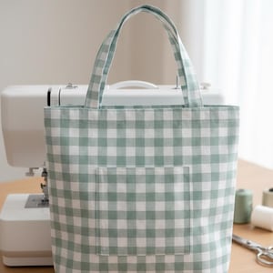 Peut inclure: Un sac fourre-tout à carreaux verts et blancs avec une poche avant et des poignées assorties. Le sac est posé sur une surface en bois, avec une machine à coudre en arrière-plan. Le sac est vert clair.
