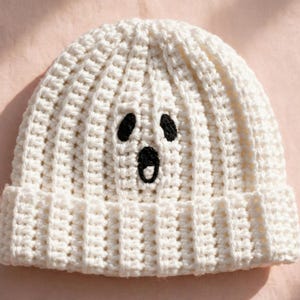 May include: A white crocheted beanie featuring a ribbed texture and a black embroidered ghost face. The design includes two oval-shaped eyes and a circular open mouth, creating a simple expression on the front of the knit headwear.