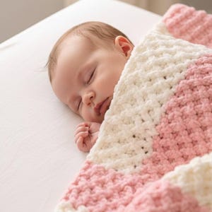 Puede incluir: Un bebé dormido está acurrucado bajo una manta de ganchillo hecha a mano. La manta presenta un patrón de puntadas texturizadas en franjas alternas de blanco y rosa suave. La piel del bebé es clara y la manta parece suave y acogedora.
