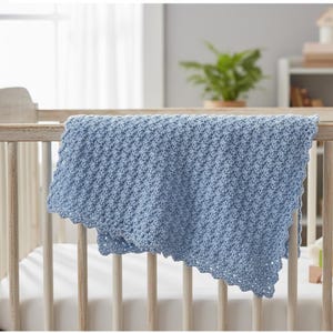 Könnte beinhalten: Hellblaue gehäkelte Babydecke, die über einem Holzbettchen drapiert ist. Die Decke hat ein strukturiertes Muster und einen gewellten Rand. Das Bettchen ist aus hellem Holz gefertigt und hat eine weiße Matratze.