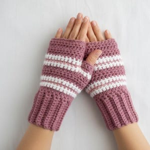 Könnte beinhalten: Ein Paar mauvefarbene Fingerhandschuhe mit weißen Streifen. Die Handschuhe sind handgehäkelt mit gerippten Bündchen und einem Streifenmuster um das Handgelenk. Auf weißem Hintergrund.