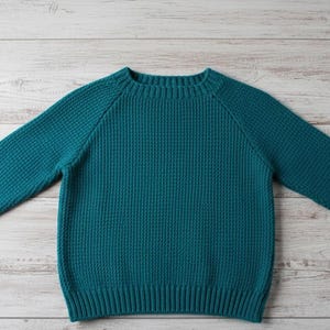Puede incluir: Jersey de color turquesa con cuello redondo y mangas largas. El tejido de punto tiene un patrón texturizado y puños y dobladillo acanalados. El jersey está tendido sobre una superficie clara de vetas de madera.