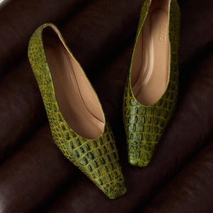 May include: A pair of olive green pointed-toe flats featuring a textured crocodile-embossed pattern. The shoes have a low-cut vamp, a smooth tan interior lining, and the letters OMG are embossed on the inner heel area of the footbed.