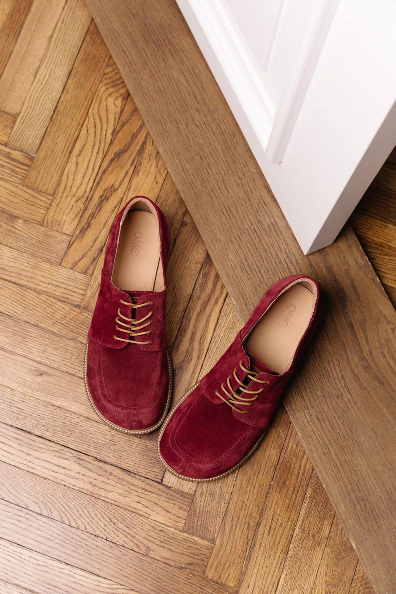 May include: A pair of deep burgundy suede lace-up shoes with tan laces and a rounded toe box. The interior lining is a light beige color with the text OMG printed on the insole. The shoes feature a visible stitched welt along the outer edge.