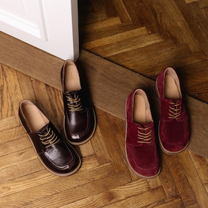 May include: Two pairs of lace-up shoes with rounded toes and light-colored stitching along the soles. One pair is crafted from smooth dark brown leather, while the other pair is made from deep burgundy suede. Both feature tan laces and tan insoles.
