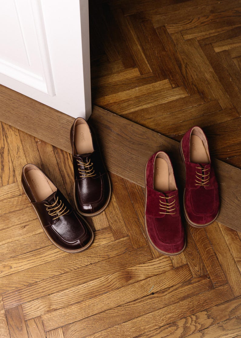 May include: Two pairs of lace-up shoes with rounded toes and light-colored stitching along the soles. One pair is crafted from smooth dark brown leather, while the other pair is made from deep burgundy suede. Both feature tan laces and tan insoles.